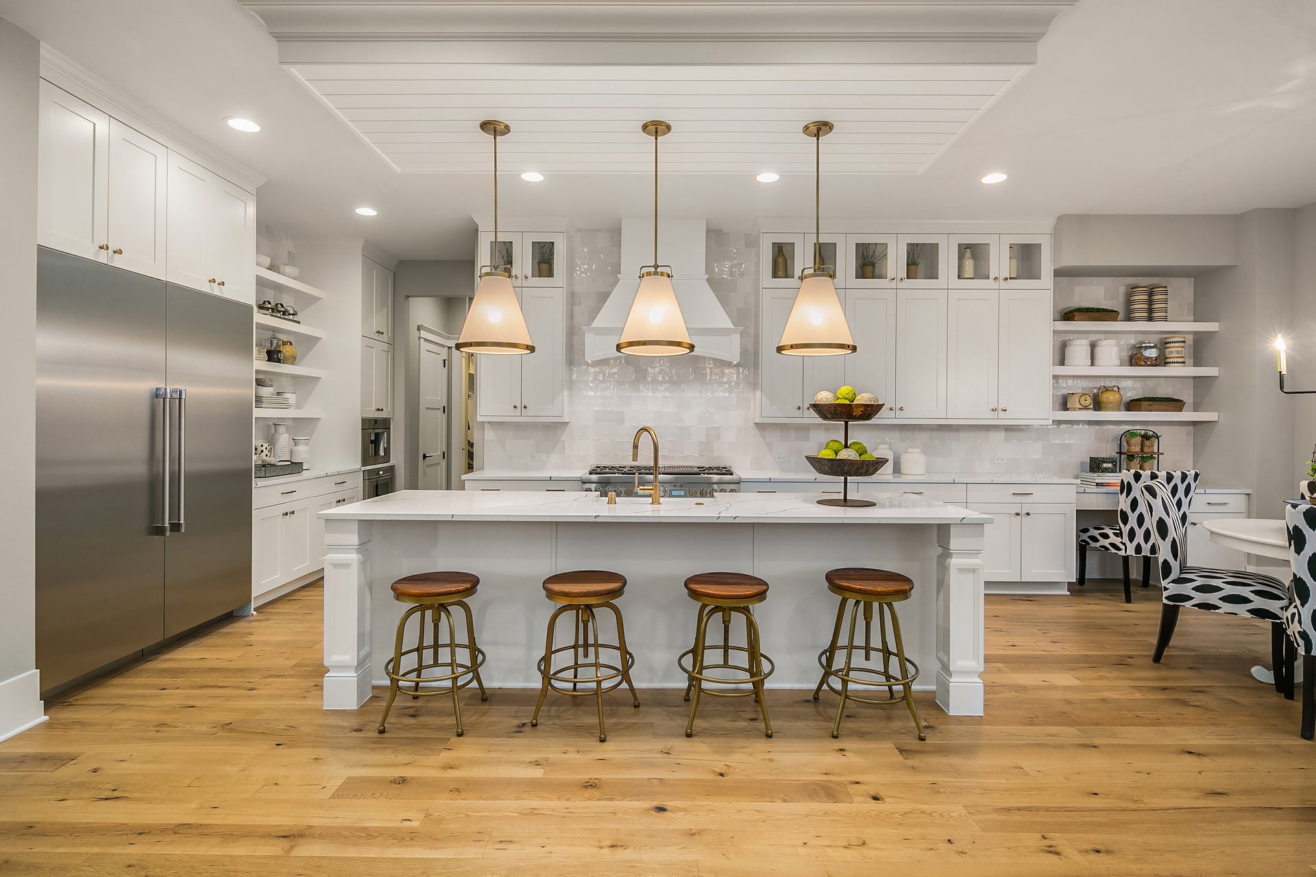 Large white kitchen with bar seating, induestrial sized fridge and gold finishes. Kitchen features an oversied island, work space, floor to ceiling white cabinets and stone countertops.